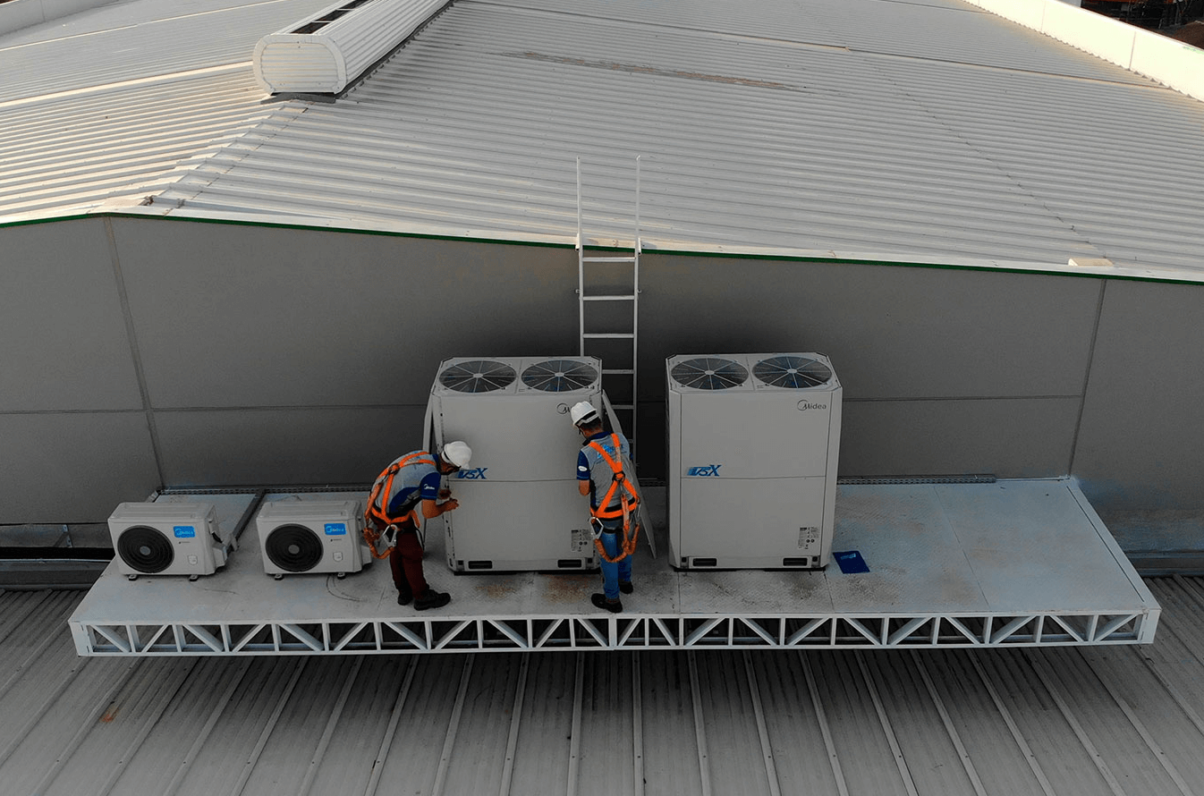 Dois colaboradores da Ambiente Ar estão no telhado finalizando a instalação do sistema VRF. Os colaboradores estão camiseta e calça e equipamentos de segurança, como corda e capacete.