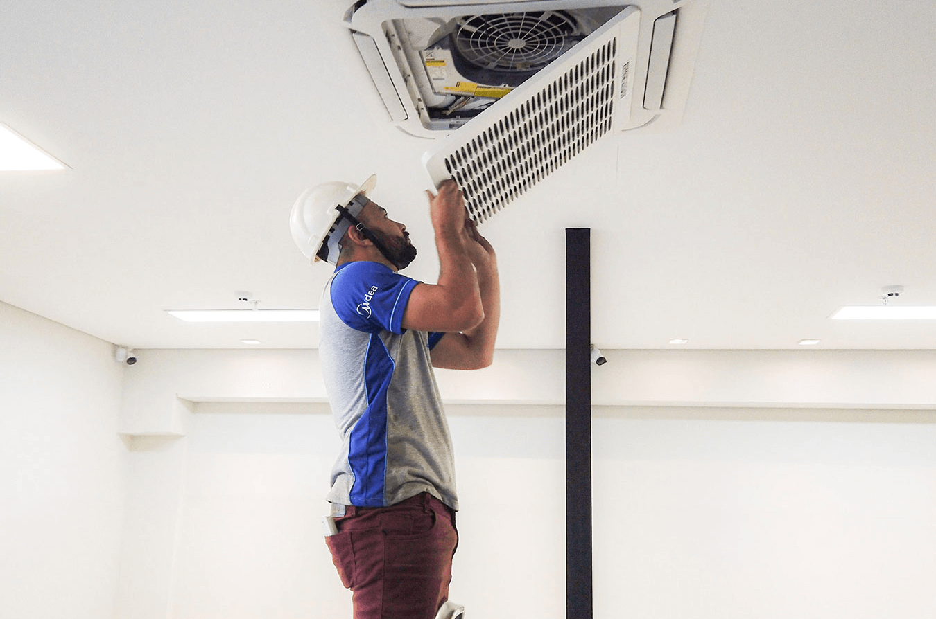 Em sala iluminada com paredes brancas, um colaborador da Ambiente Ar está instalando o ar-condicionado VRF no texto. O colaborador veste a camiseta do uniforme, uma calça bordô e capacete de segurança. 